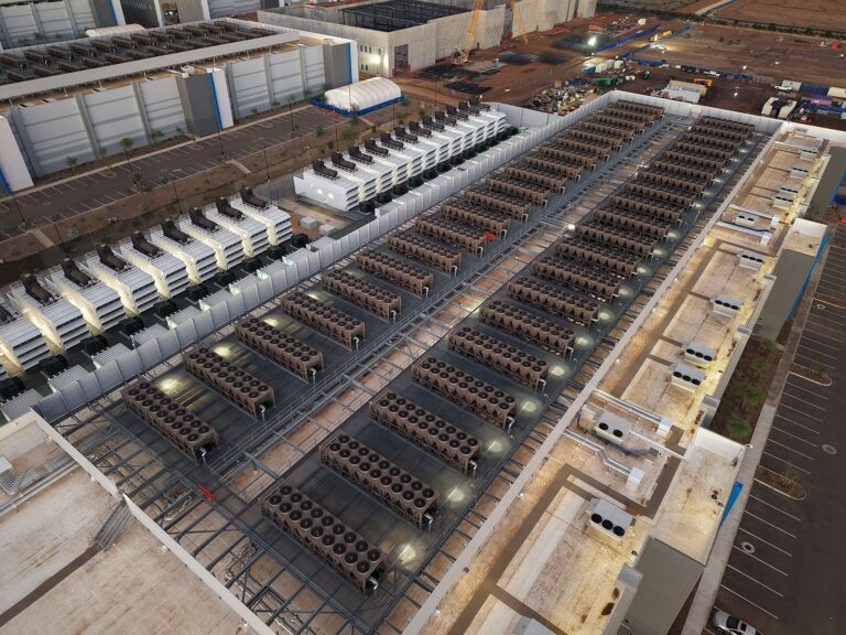 Aerial view of a data center roof lined with rows of cooling units and backup generators, with more data center buildings under construction behind it