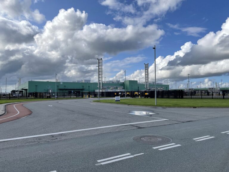Long green data center buildings behind a security fence, seen across an empty road under a cloudy blue sky