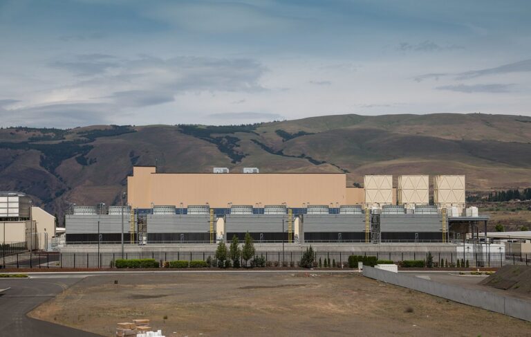 Large tan data center building with rows of cooling units in front of brown hills under a cloudy sky