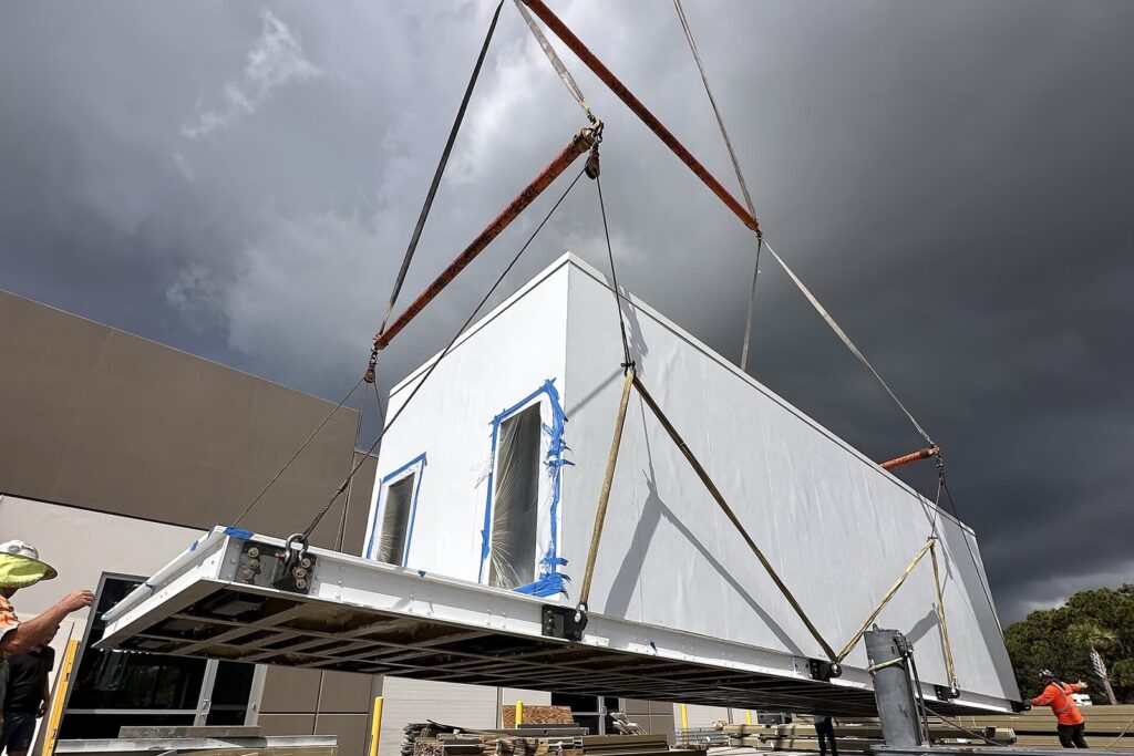 A crane lifts a white prefabricated modular data center unit off a truck at a commercial building site