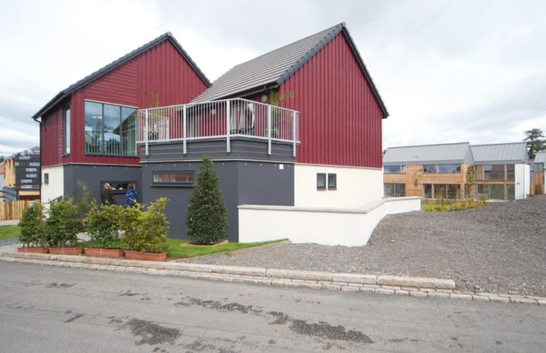Finished two-storey modular house with red timber cladding and a balcony, beside other modular homes